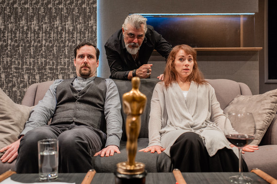 Two people sit on a sofa looking at an Oscar statue on the coffee table in front of them. They look equally stunned and fearful of the statue. Behind them, a man leans on the couch looking at the award with great pride.