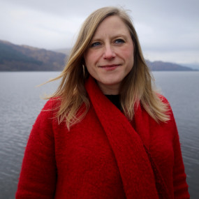Frances Poet has long blonde hair. She is wearing a red coat and standing in front of a lake.