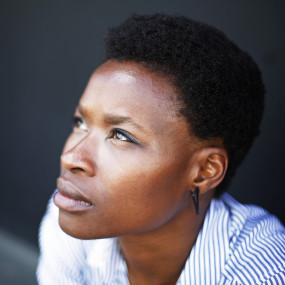 Adura Onashile as a short, black afro hairstyle. She is wearing a blue striped shirt,